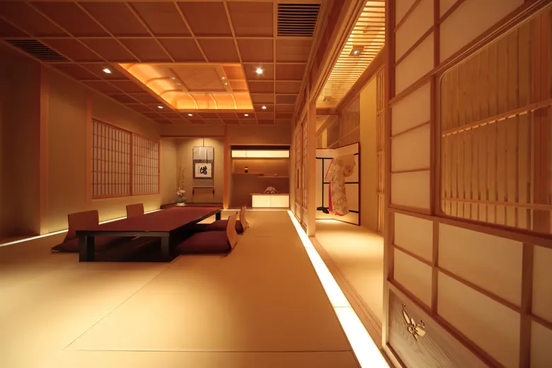 experiences_kyoto_private-geisha-entertainment-dinner-gion-kyoto_ochaya-man-teahouse-interior-gion-higashi The serene, softly illuminated interior of a private ochaya tatami dining room in Gion-Higashi, Kyoto, featuring low wooden banquet tables with floor cushions, shoji sliding screens, a hanging scroll, and a displayed kimono.