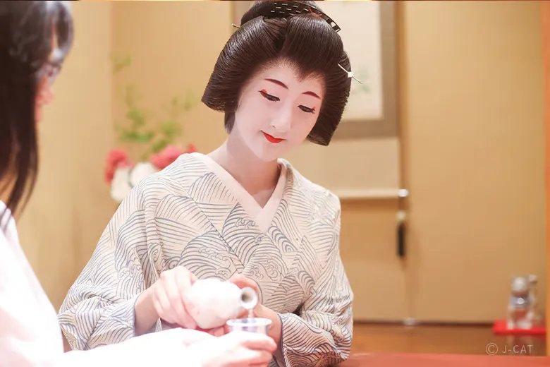 experiences_kyoto_private-geisha-entertainment-dinner-gion-kyoto_pouring-sake-for-guest A senior geiko in traditional makeup and silk kimono smiling as she gracefully pours sake from a ceramic tokkuri carafe into a guest's cup during an intimate private geisha dinner in Gion, Kyoto.