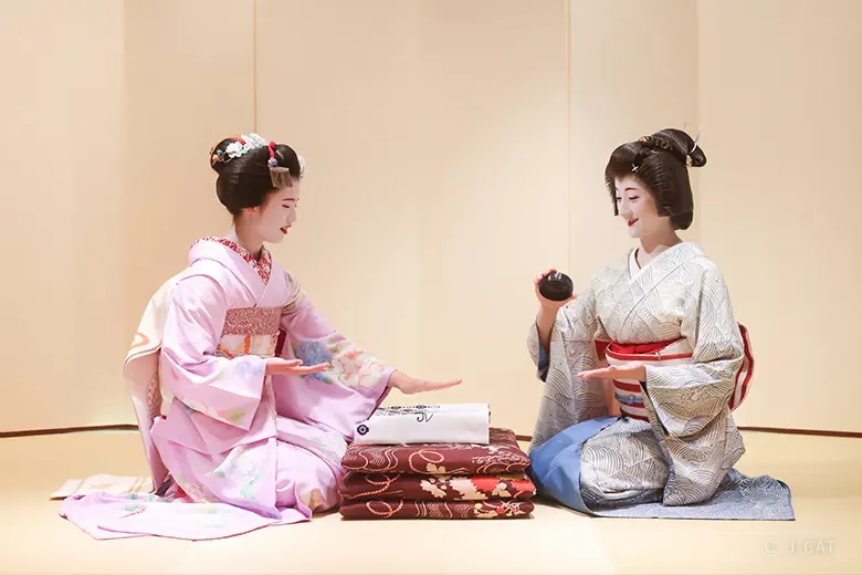 An apprentice maiko in a light pink floral kimono and a senior geiko in a patterned white kimono sitting on tatami mats during a private geisha entertainment dinner in Gion, Kyoto, engaging in a traditional ozashiki asobi parlor game.